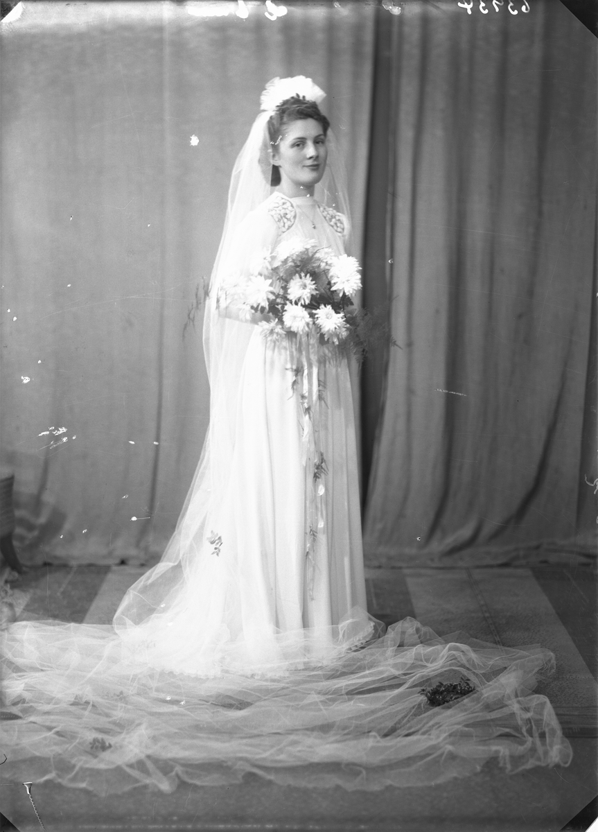 Portrett. Brudebilde. Ung mørkhåret kvinne i lang lys brudekjole med pynt på skuldrene, slør og blomsterbukett. Brud. Bestilt av Hr. Henrik Styve.
