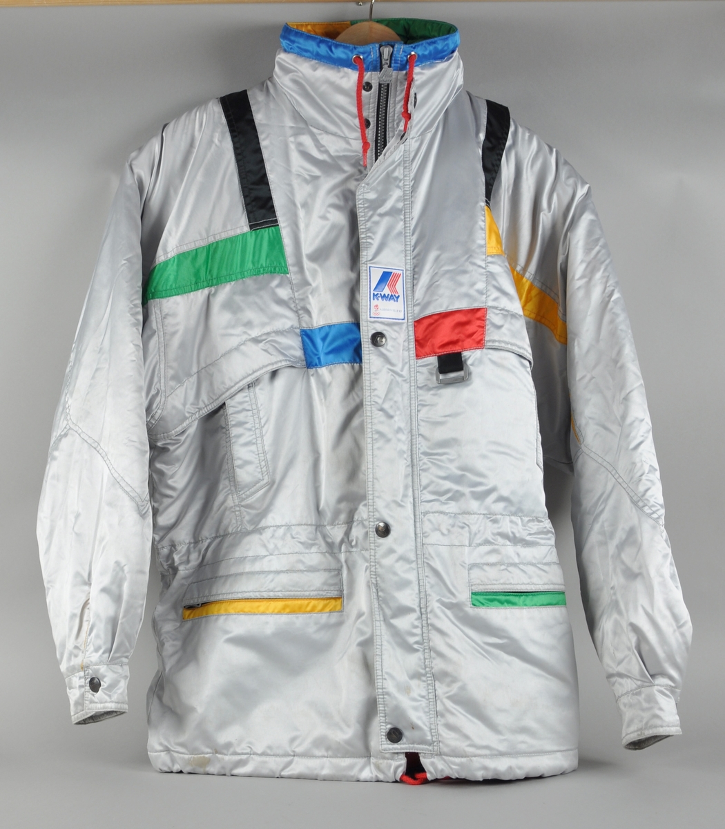 Grå jakke med dekor i ulike farger blant annet ved lommer og krage. Påsydd merker med logo for K-WAY og emblemet for de olympiske vinterleker i Albertville i 1992.