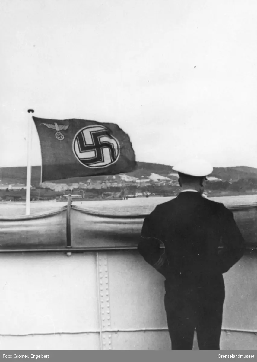 Skipsoffiser står akterut på et skip som går fra land. Flagg med hakekors som motiv med tysk ørn stående på hakekors i øvre hjørne ved festeanordning. 