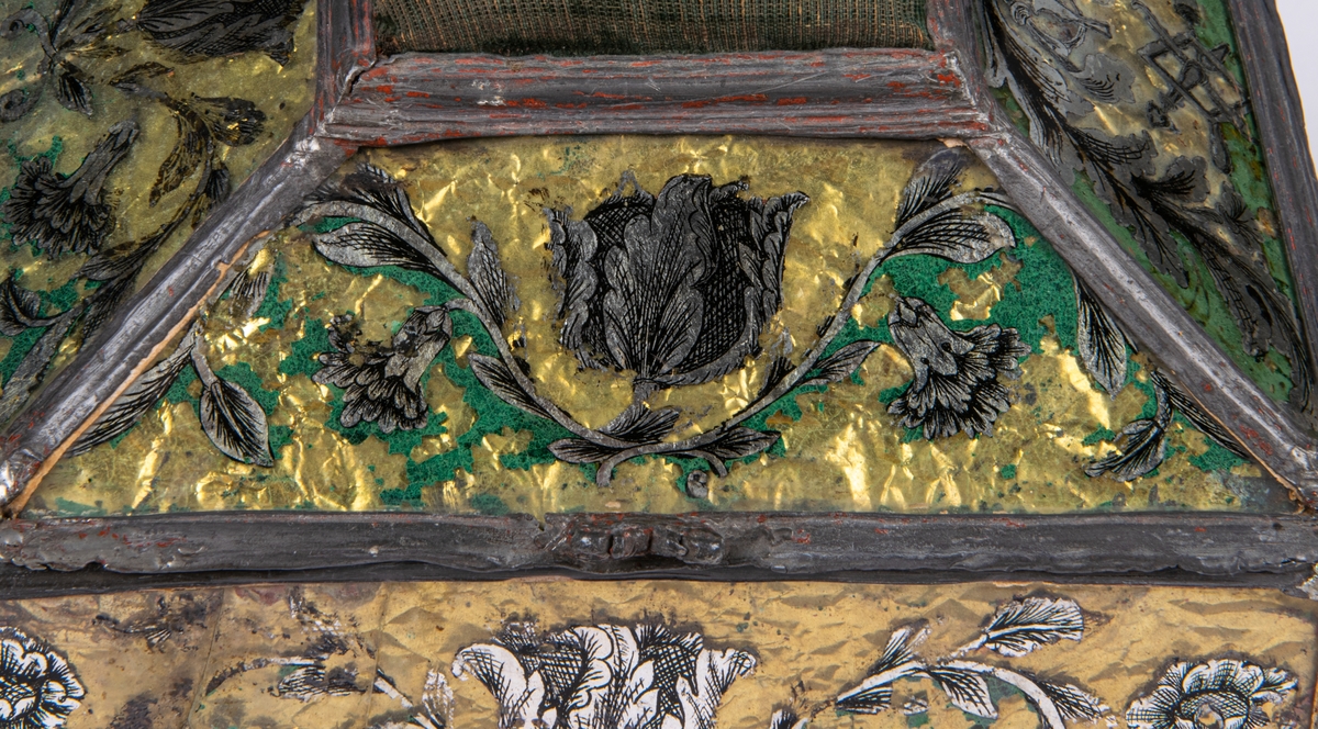 Syskrin. Överst grön nåldyna. Formen är pulpetartad, sidorna bestå av målade glasskivor med blylister emellan. Målningen är grön med blomornament i svart och silver. Gult papper under. Locket rör sig på två gångjärn och hålles innuti av en tunn kedja. I locket under nåldynan finnes en liten spegel med målning i grönt "W.E.M. 1767", med kunglig krona över samt växtranka omkring nertill. Skrinet står på fyra små kulfötter av bly. På ena fältet av locket ser man glasmästareämbetets sigill. Gävle, Gästrikland.