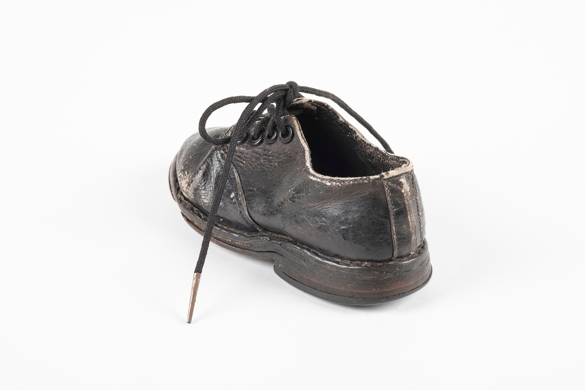 Et randsydd barnesko (venstre sko) av lær. Skoen har snøring med runde lisser. Maljene er av metall. Sålen er av lær og er forsterket med spiker på hælen og ved tuppen på undersiden.