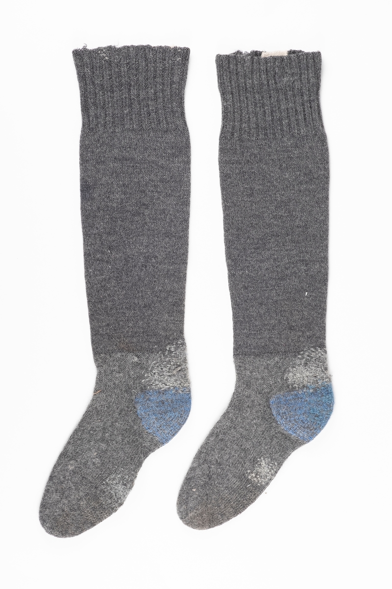 Ett par knestrømper strikket med grått ullgarn. Hælen er strikket med blått garn. På innsiden ved åpningen i begge strømper er det påsydd en lapp med fangenummer.