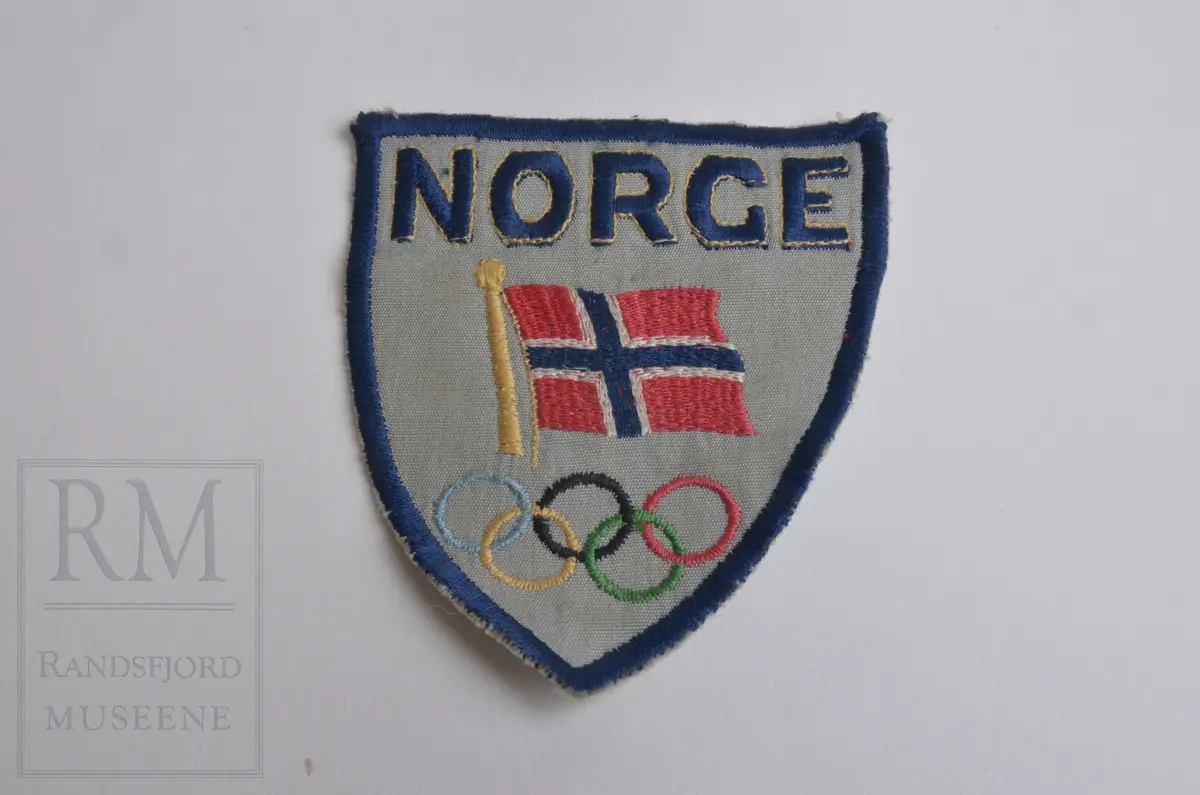 Grått stoff fôret med hvitt stoff bak. Brodert blå kant, teksten NORGE, det norske flagget og OL-ringene. Maskinsøm.