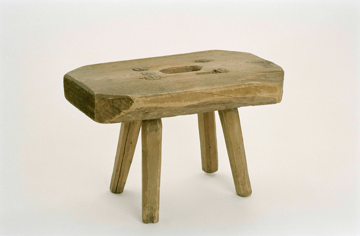 Pall, mjölkstol, av furu. Rektangulär sits med avskurna hörn, ovalt handgrepp mitt på sitsen. Fyra ben, något utställda.