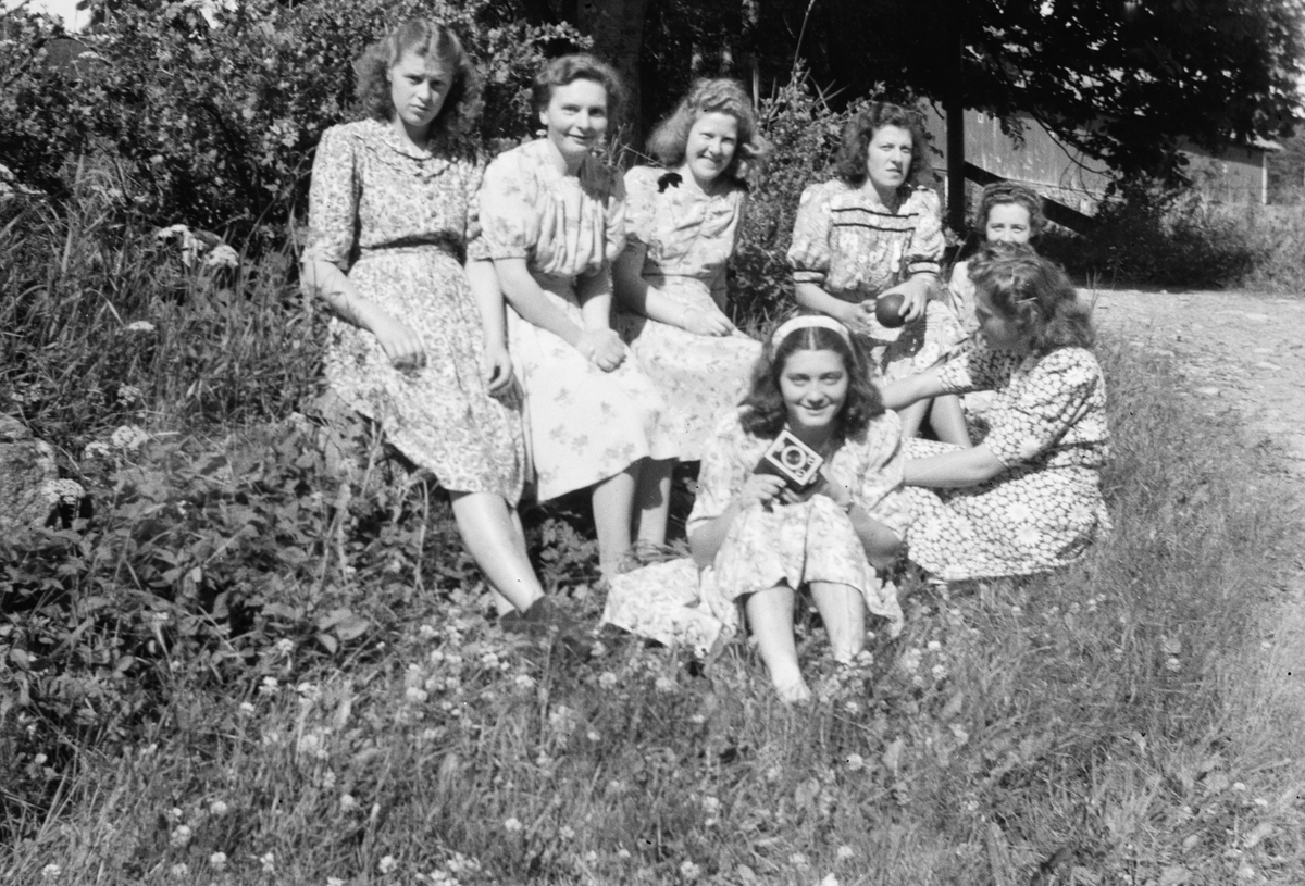 Gruppefotografi av sju unge kvinner i kjole. Den fremste kvinna har et kamera? i handa.