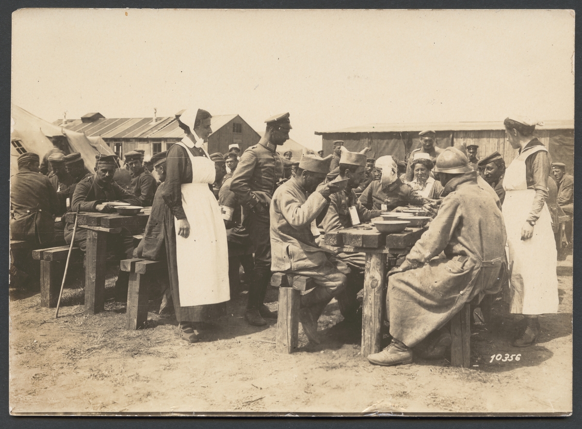 Bilden visar franska krigsfångar och sårade som sitter vid grov snickrade bord och äter middag. Flera soldater bevaka de och två kvinnor med vita förkläden talar med de. I bakgrunden syns flera baracker och en tält.
