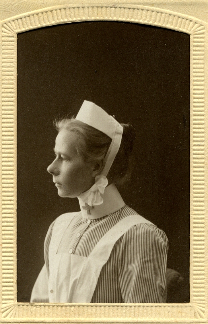 Foto av en kvinna i sjuksköterskeuniform med hätta. 
Bröstbild, profil. Ateljéfoto.