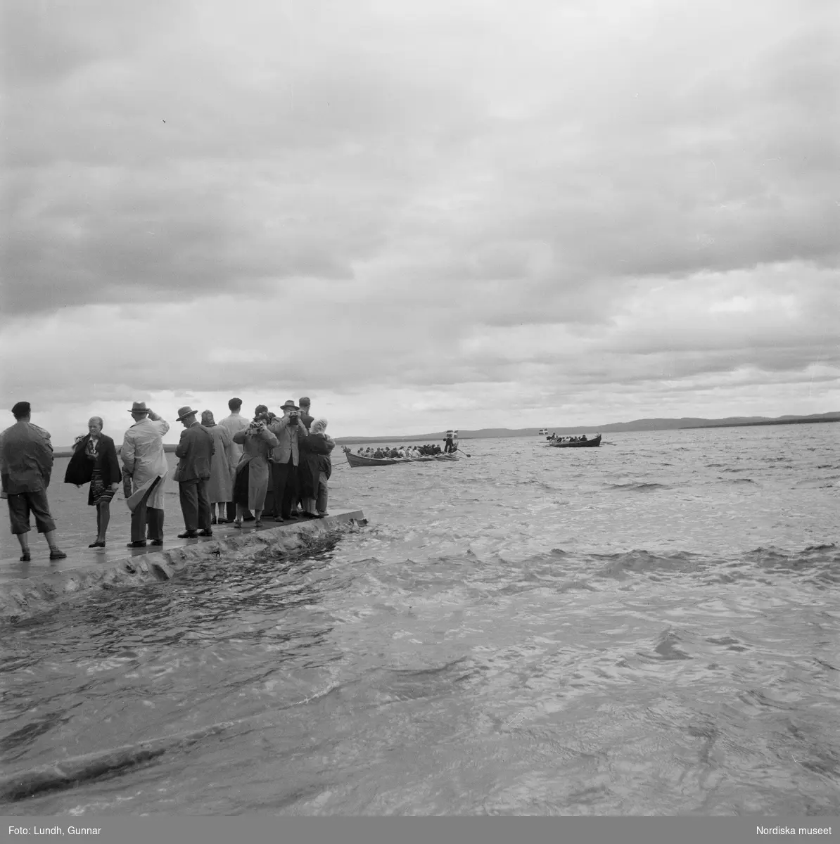 Motiv: (ingen anteckning) ;
En folksamling står vid en brygga med två kyrkbåtar, kvinnor och män ror kyrkbåtar i samband med midsommarfirande.