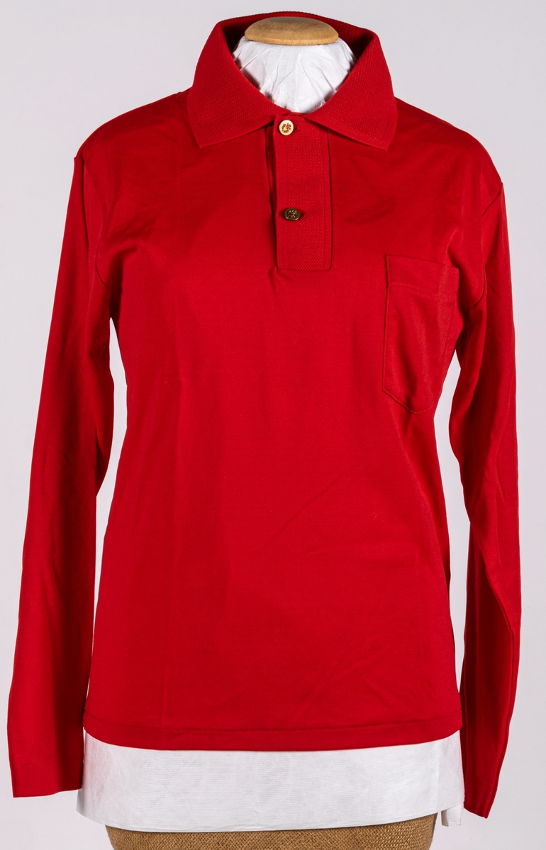 Röd, långärmad tröja av banlon.
En bröstficka, krage och sprund fram försedd med två metallknappar.
Runt översta knappen hänger en papperslapp med tillverkarns namn och priset: 32:75.
Konfektionssydd.