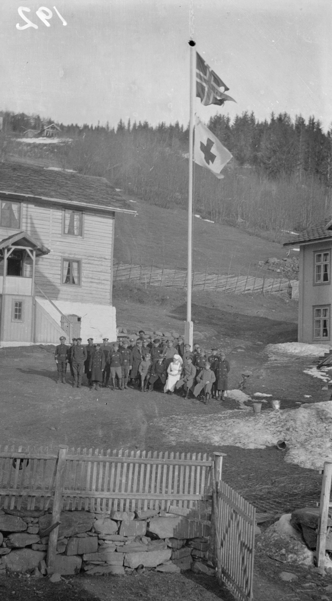 Gårdstun med flaggstang med norsk splittflagg og Røde Kors-flagg, mange mennesker oppstilt - internerte under første verdenskrig?
