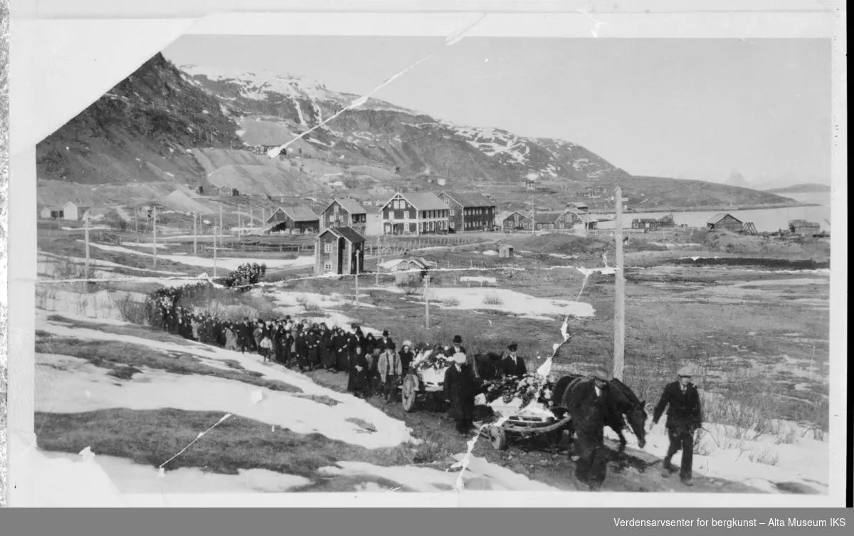 Begravelsesfølge til Hoel og Erntsen i Kåfjord med flere hus og fjell i bakgrunn.