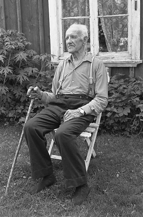 En äldre man, Karl Fransson, sitter i en trädgårdsstol framför ett hus med locklistpanel. Han är klädd i skjorta och byxor med hängslen och strumpor utan skor. Han håller en käpp i höger hand.