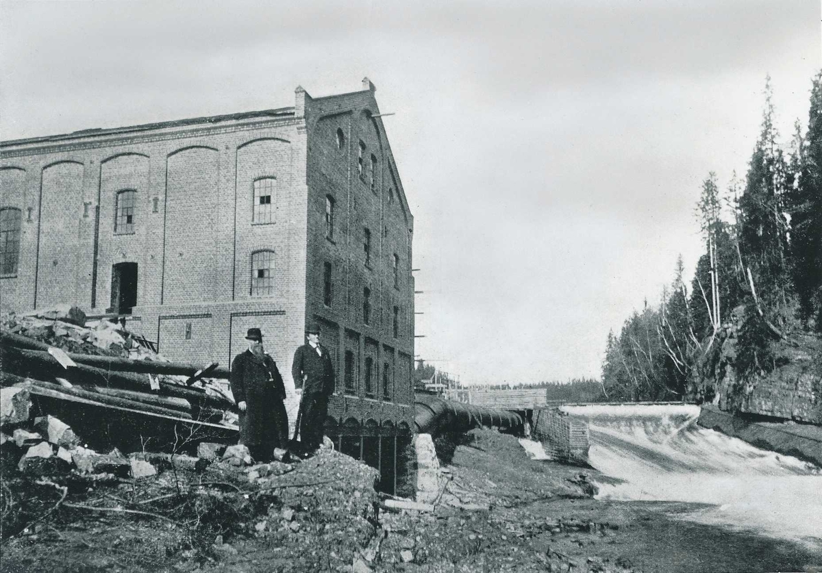 To menn står foran en industribygning i teglstein. I bakgrunnen er en foss med demning og daminretning. 