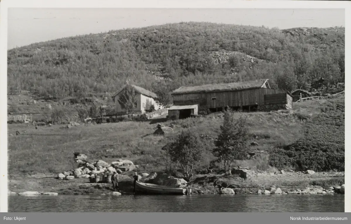 Gården Skinnarland sett fra innsjøen Møsvatn. Ved vannkanten står to personer ved personbåt. I lua sees bolighus, låve og ulike driftsbygninger