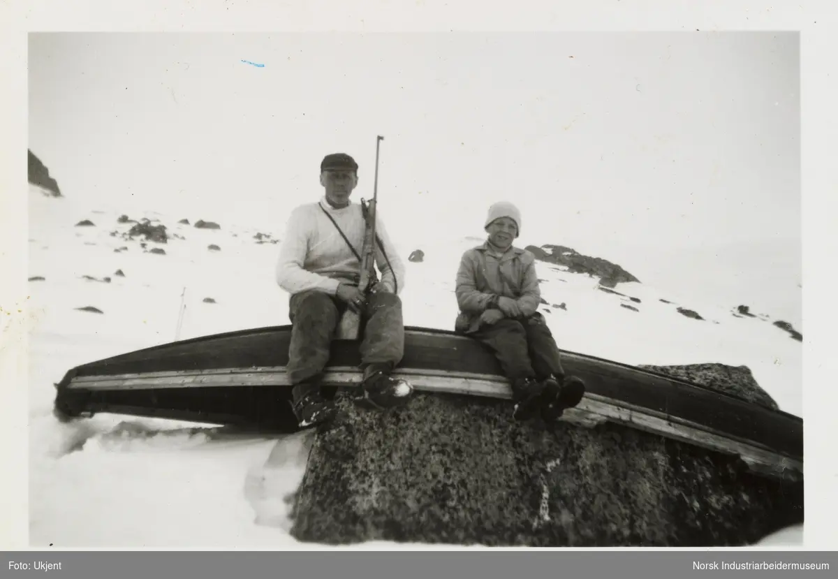 Olav Vågen holder gevær og sitter på en pråm sammen med sønnen Johan. Pråmen ligger opp ned på en stein i snøen på Tuddeltjønni, Møsstrond