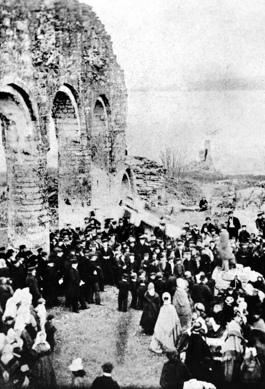 Kornete svart-hvitt foto av en stor menneskemengde i penklær under de runde buene til domkirkeruinen på Domkirkeodden.