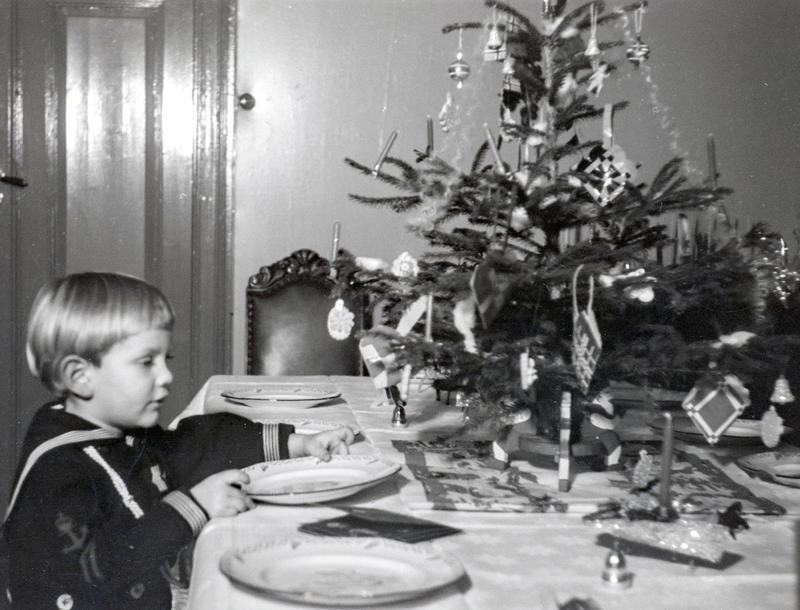 Svart-hvitt foto av en liten gutt i matrosdress som dekker bordet. På bordet står også et lite juletre.