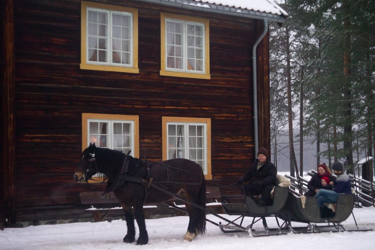 Hest og sluffe foran Gjersøyen-bygget. Brun hest. Snø på bakken. Fotografiet er tatt under arrangementet Jul i stuene. I sluffa sitter en barnefamilie.