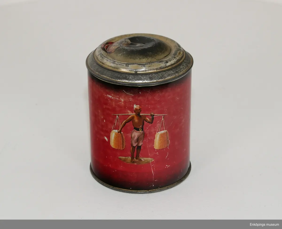 Röd plåtburk med lock, märkt på ovansidan "2 Kaffe!". Har en målad figur bärande på ett ok.