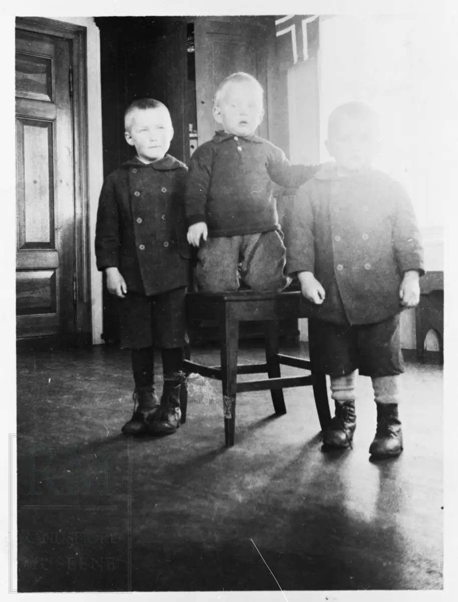Tre gutter, brødre på Gisleberg, står oppstilt i et rom. Et norsk flagg ses i bakgrunnen.