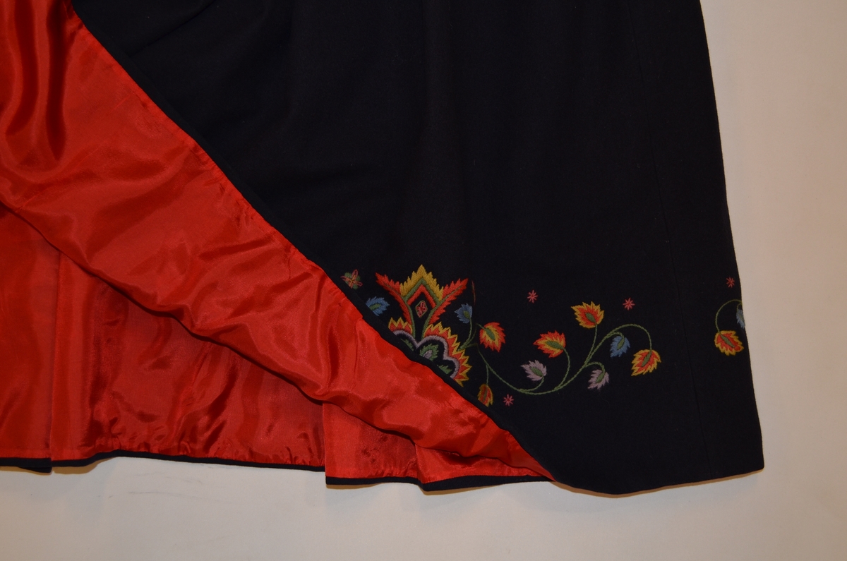 Valdresbunad (S1) i svart ulltøy med kulørt ullbroderi. Stakk A, lomme B og lue C. Stakken er fòra med 20,5 cm raudt silkefór på innsida av stakken.