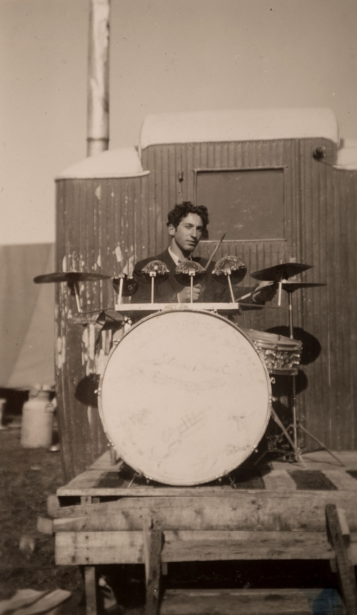 En ung romsk man spelar trummor framför en vagn, Sandviken juni 1947. I bakgrunden syns en skorsten samt upphängda tältdukar i ett läger.