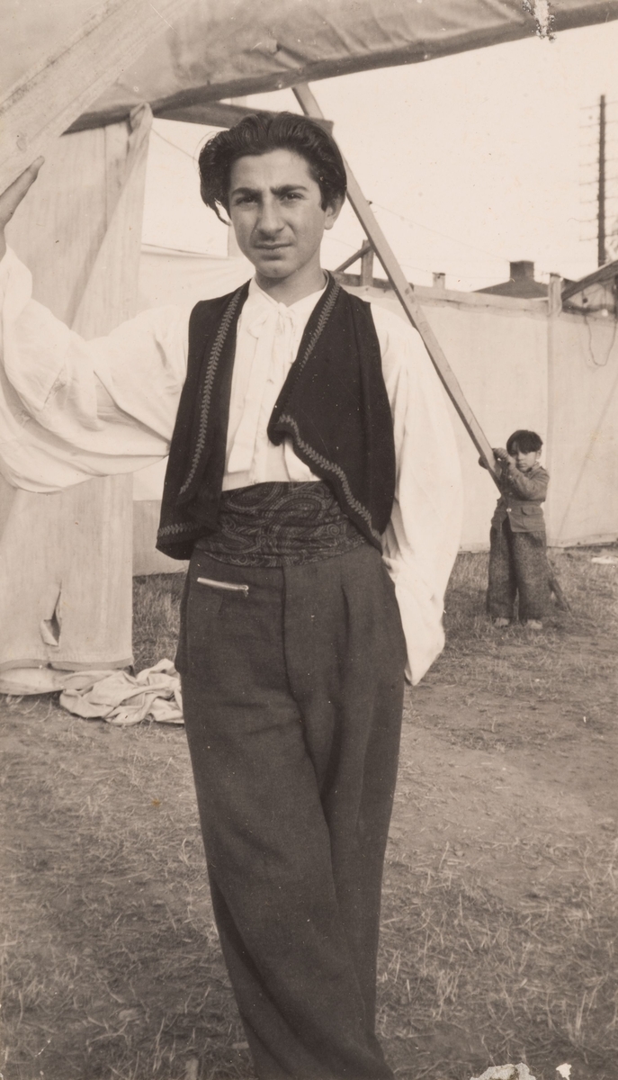 En romsk pojke står i ett läger. I bakgrunden syns en mindre pojke och tält. Tältdukarna var ofta uppspända för att avskärma lägret och hålla obehöriga utanför. Fotografiet är antagligen taget i trakterna runt Sandviken sannolikt år 1947.