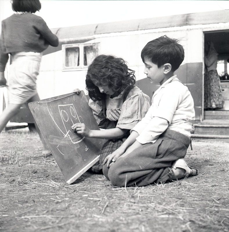 Fotografiet är taget i juli 1952 i samband med skolundervisning i det romska lägret i Ekeby, ca 4 km väster om Eskilstuna. Två barn sitter på marken och ena barnet ritar med en krita på en griffeltavla.