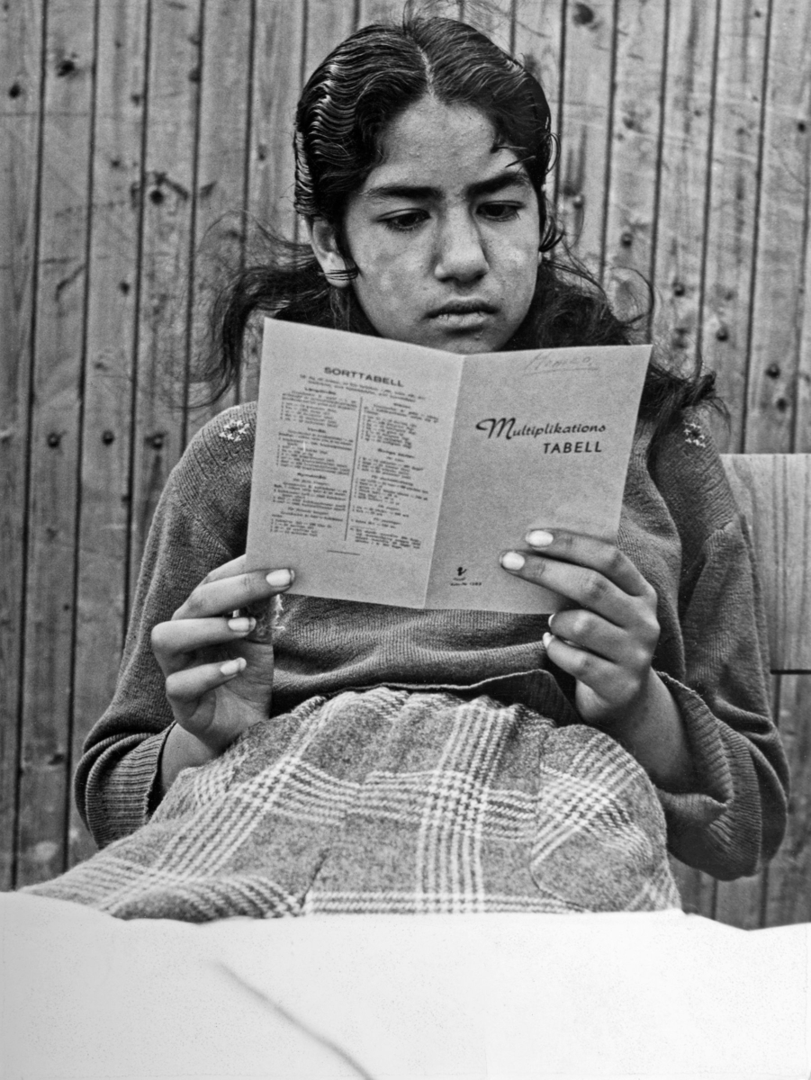 En romsk flicka läser en multiplikationstabell under lägerskolan 1954. Hon sitter lutad mot en trävägg av något slag.