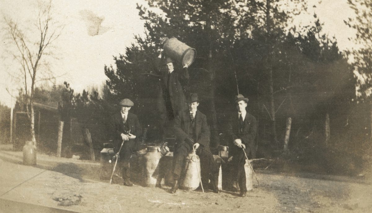 Portrettfotografi av fire unge menn med hatt, frakk og en pinne i handa. De sitter på noen melkespann på en vei.