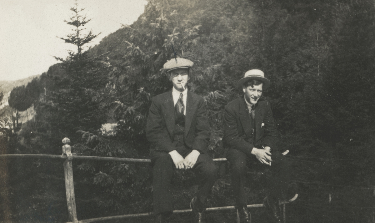 Portrettfotografi av to unge menn i dress som sitter på et gjerde ved en skog. Mannen til venstre er identifisert som Tøssebro.