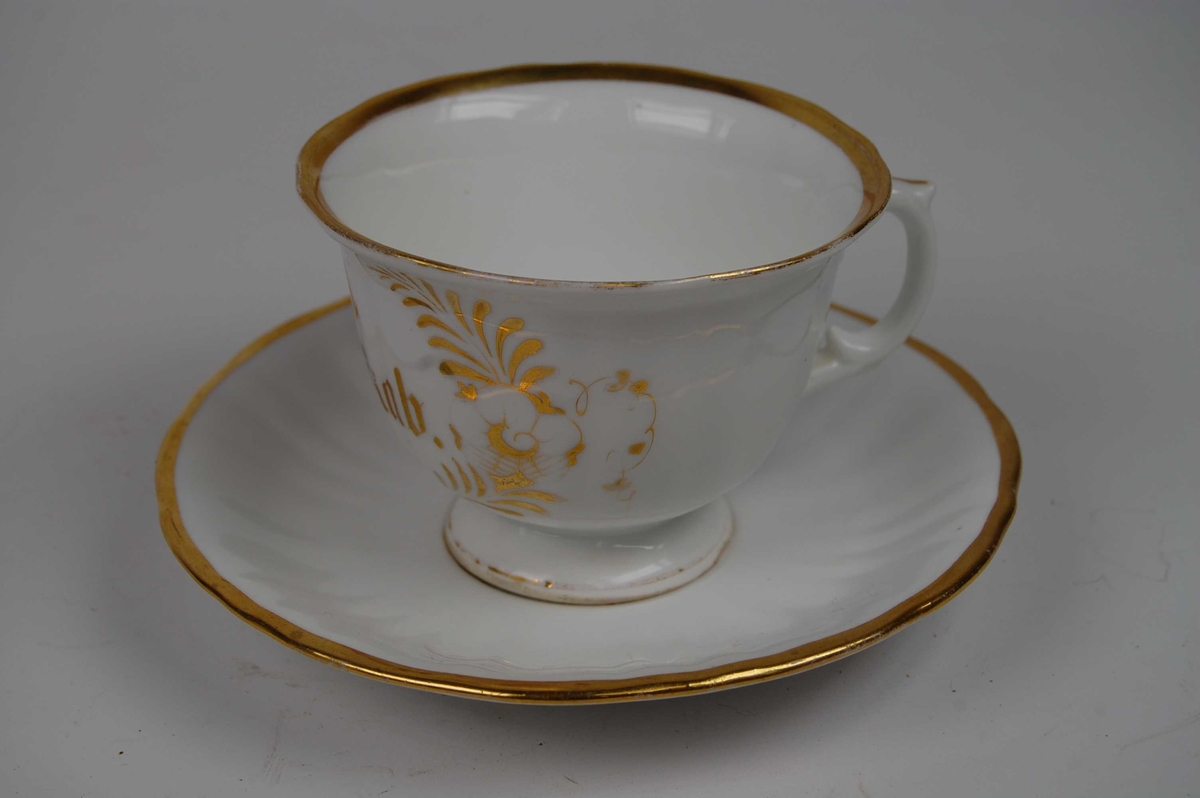 Kaffekopp med hank og skål. Gullkant langs munningen og på skålen. Blomsterranker trykt i gull danner en kartusj som gir rom for tekst. 