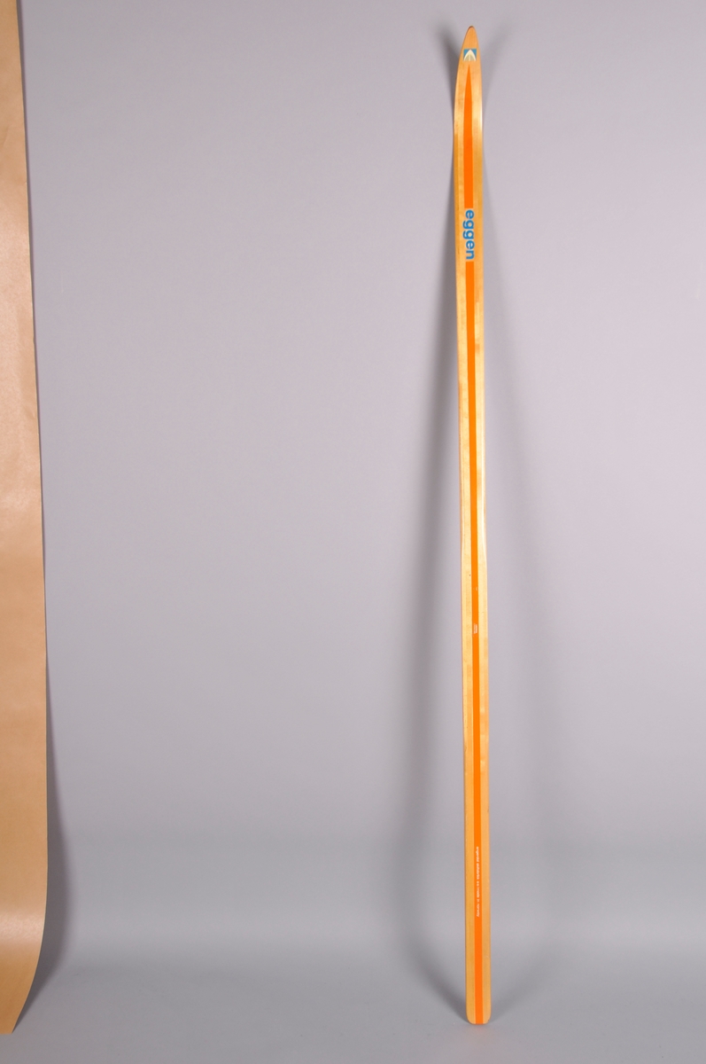 Langrennski i tre, lakkera trekvit overflate, oransje stripe på overside og på sidekantar. Emblem på skitupp. 