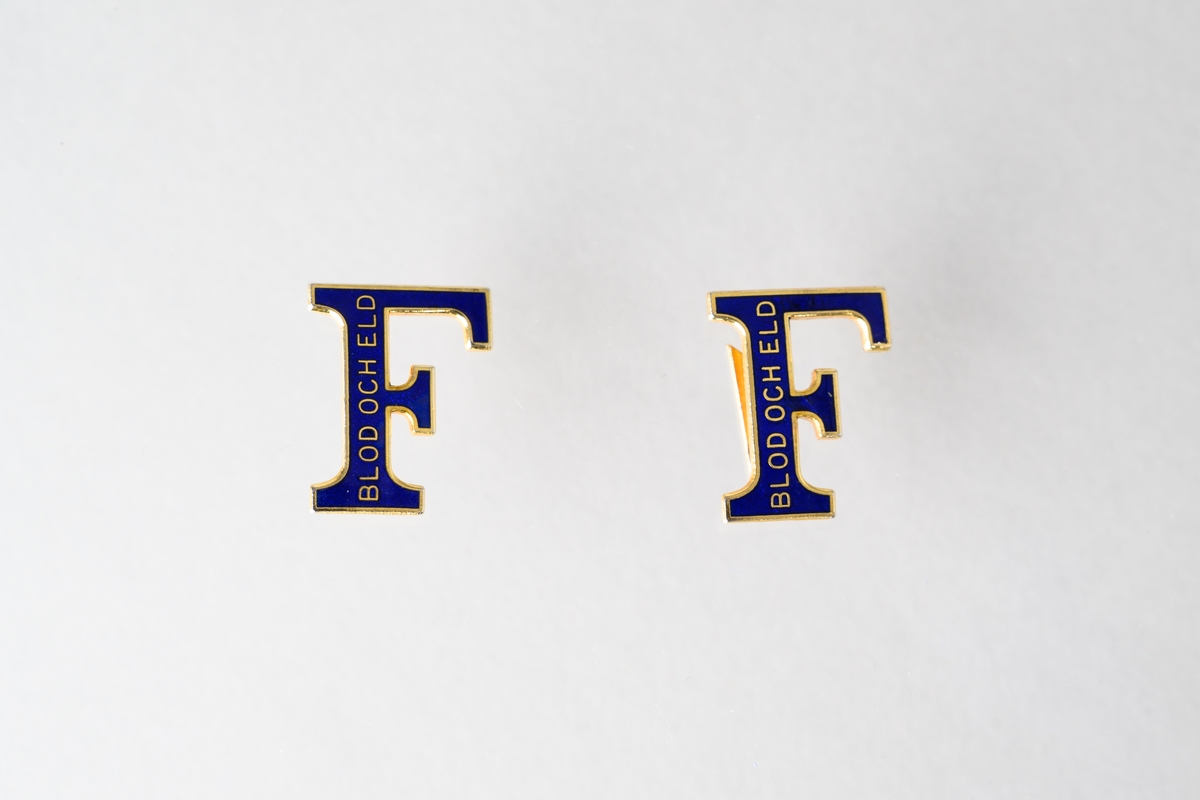Märke, 2 st, av metall i form av ett "F", för rockslag till uniformskavaj för kvinnlig soldat i Frälsningsarmén. Märket är i färgerna blått och guld med text: "BLOD OCH ELD". Märkerna hör till rock JM.56618:1

Det vänstra märket (när man har kavajen på sig) är instucket uppifrån på litet sexkantigt, blått tyg (labell) och fastsytt på tyget, som i sin tur är fastsytt på rockslaget. Det högra märket är instucket underifrån på litet sexkantigt, blått tyg (labell) och fastsytt på tyget, som i sin tur är fastsytt på rockslaget.
