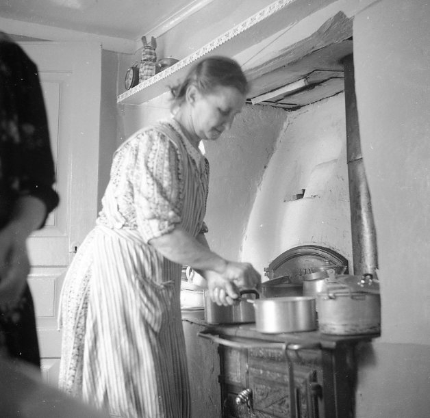 Kvinnor (2) som lagar mat vid en vedspis.