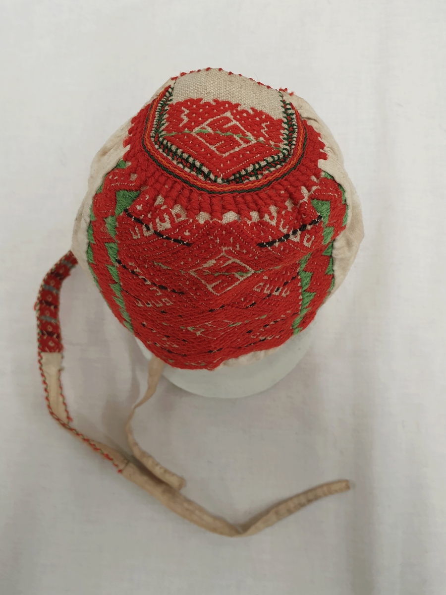 Barnekyse av hvit bomull med brodering i rødt, grønt og svart på bakhodet. Bånd med brodering på innsiden av båndet.