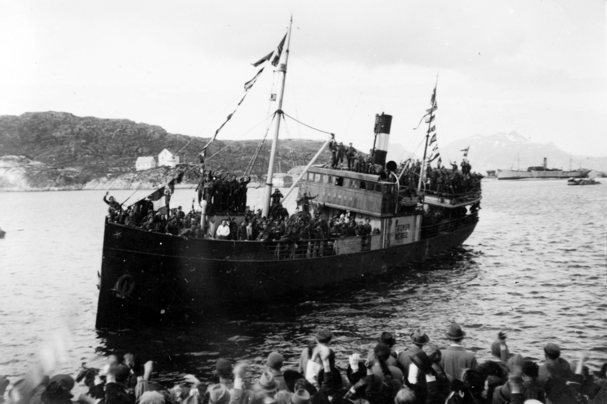 D/S "Tromsø" ankommer en havn. Ombord er det soldater, i forgrunnen tilskuere på kaia.