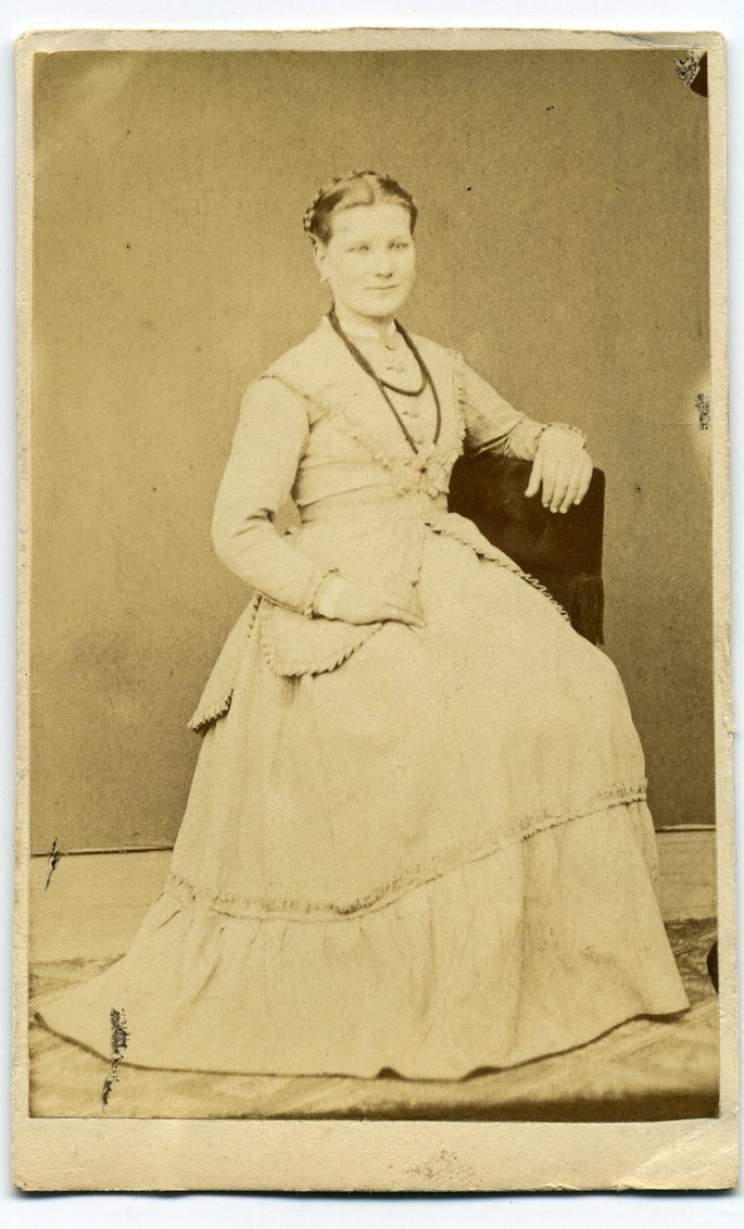 Kabinettsfotografi: porträtt av okänd ung kvinna i ljus klänning och halsband. Hon sitter på snedden på en stol.