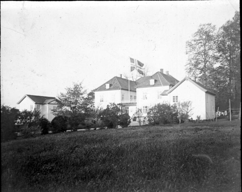 Et veldig tidlig svart/hvitt fotografi av Eidsvollsbygnignen sett på litt avstand. Et norsk flagg vaier i vinden
