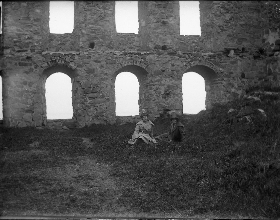 Två unga kvinnar sitter på marken i Brahehus slottsruin. De bär kappa och hatt. Det är möjligen syskonen Anna-Lisa och Karin Rythén