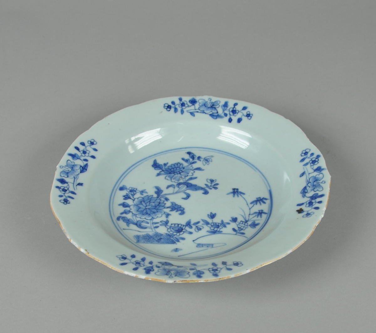 Dyptallerken av glassert keramikk. Tallerkene har bølgete rand. Det er blå dekormaling av naturlandskap og blomster på tallerkenen.