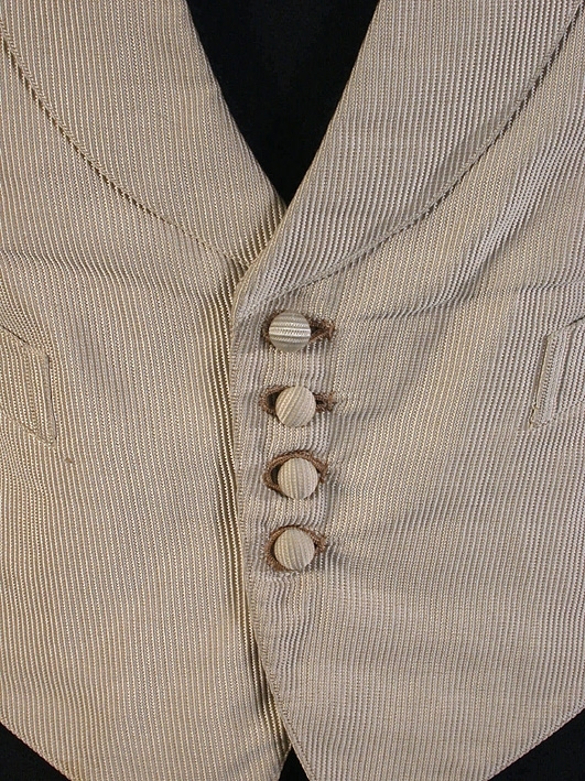Framstycket i gråbeige rip, silke i varp och bomull i inslag. Bakstycke i kyperttyg. Schalkrage. Enkelknäpp med fyra stycken överklädda knappar.