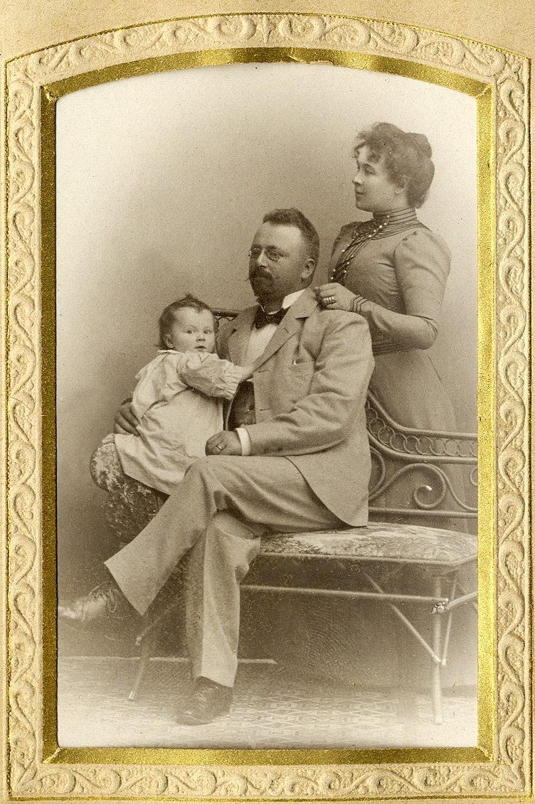 En man i ljus kavajkostym, hans fru och deras lilla dotter. Trävaruhandlare Edvard Andersson (1875-1934) med sin fru, fotografen Betty Larson (1864-1939) och deras dotter Kristina (Stina) (1899-1965). 
Helfigur. Ateljéfoto.