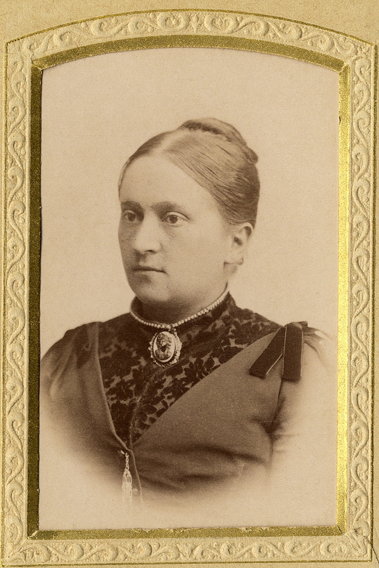 En okänd medelålders kvinna i mörk klänning med sammetsbrokad och hög krage. Vid kragen syns ett pärlhalsband och en kamébrosch. 
Bröstbild, halvprofil. Ateljéfoto.