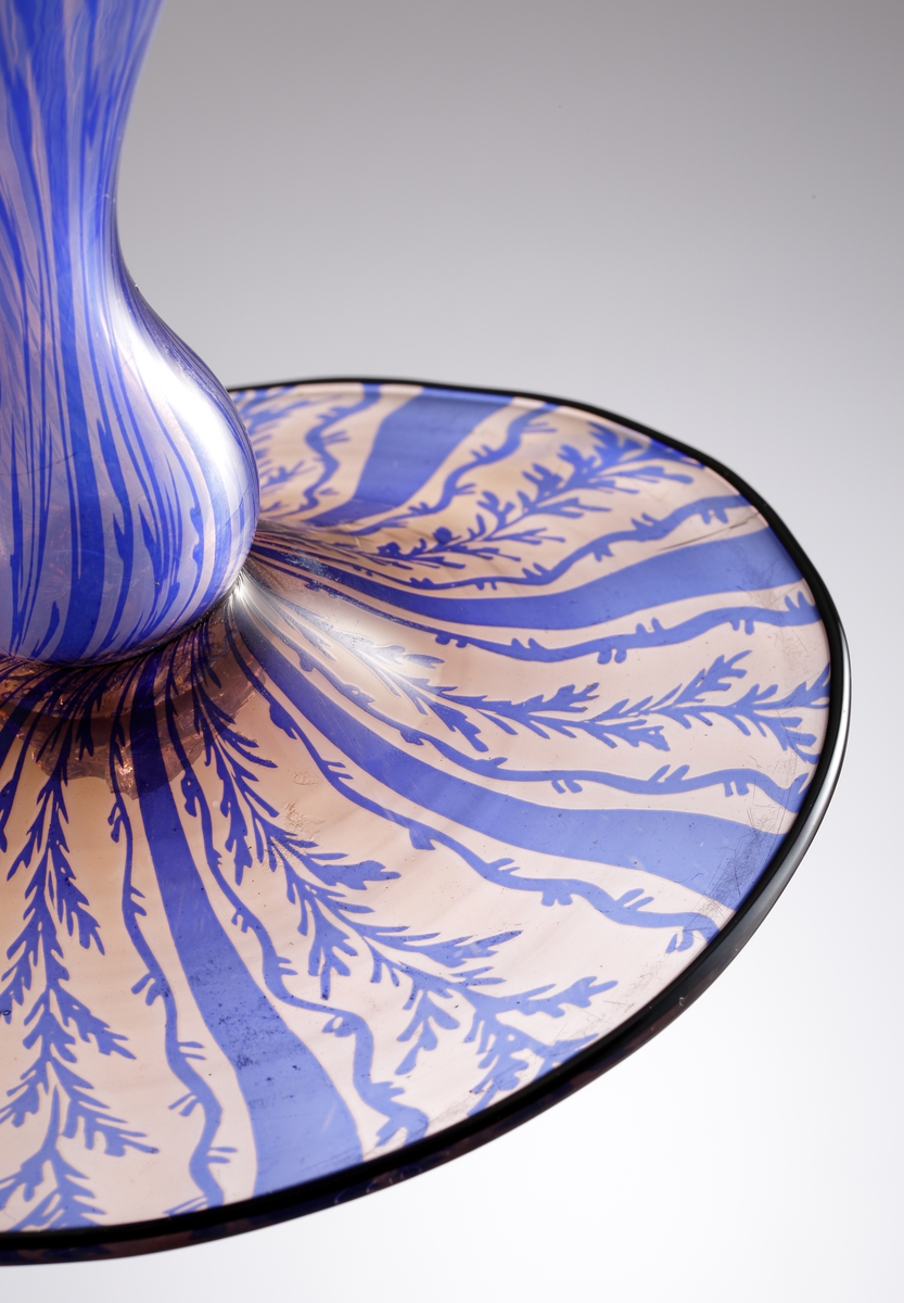 Hög graalvas, troligen Edward Hald. Vas på fot med blått växtmotiv mot ofärgad botten. Vasen är osignerad men stilen och mönstret gör gällande att den mest trolige formgivaren är Edward Hald.