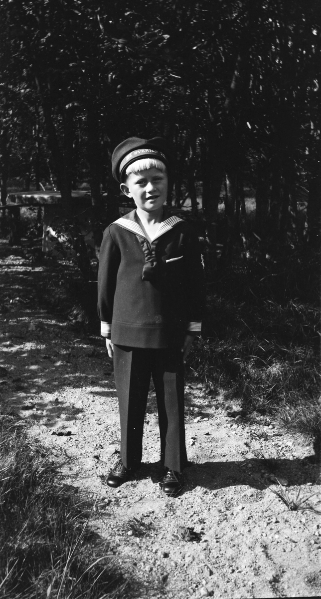 En liten pojke står i sjömanskostym och finskor på en sandad gång i en park eller trädgård. På mösskärmen står det Sverige och byxorna har skarpa pressveck.