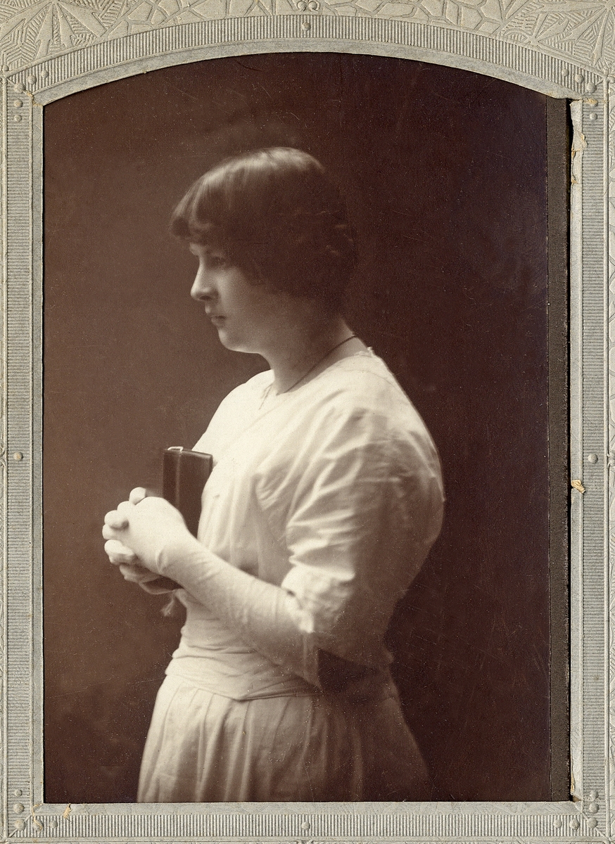 En tonårsflicka i vit konfirmationsklänning med vita handskar Hon håller i en psalmbok (?). 
Midjebild, profil. Ateljéfoto.

Fotografens dotter.