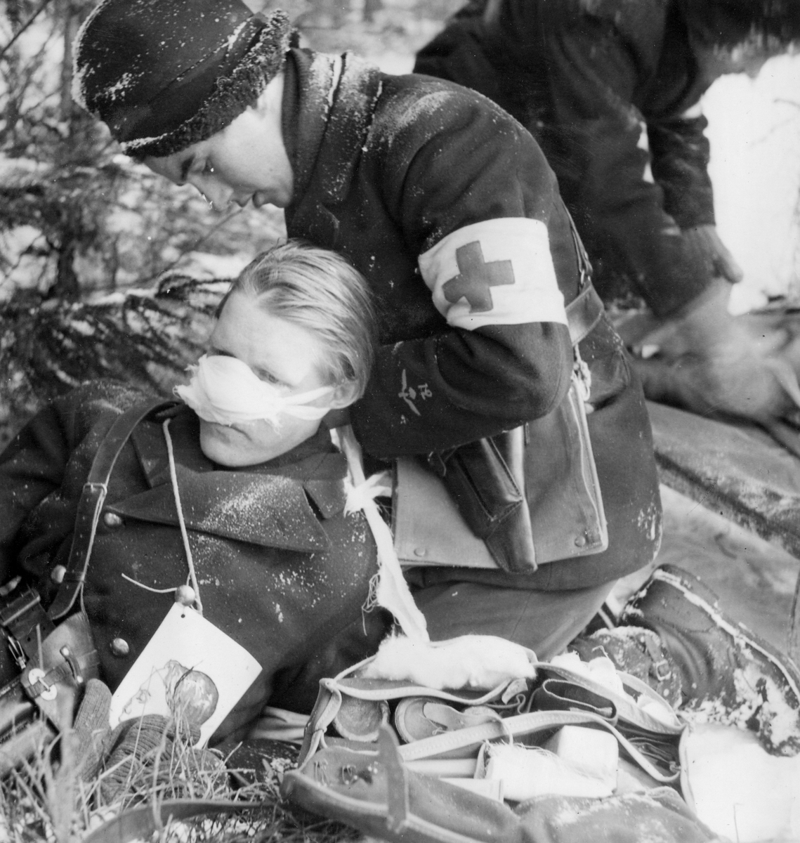 Första hjälpen utförs på soldat som agerar skadad vid sjukvårdsövning i fält vid F 11 Södermanlands flygflottilj, 1945. Omplåstring av grantskadad näsa.

Vintertid.

Ur fotoalbum "Sjukvårdsskolan 15/1-15/3 1945" från F 11.