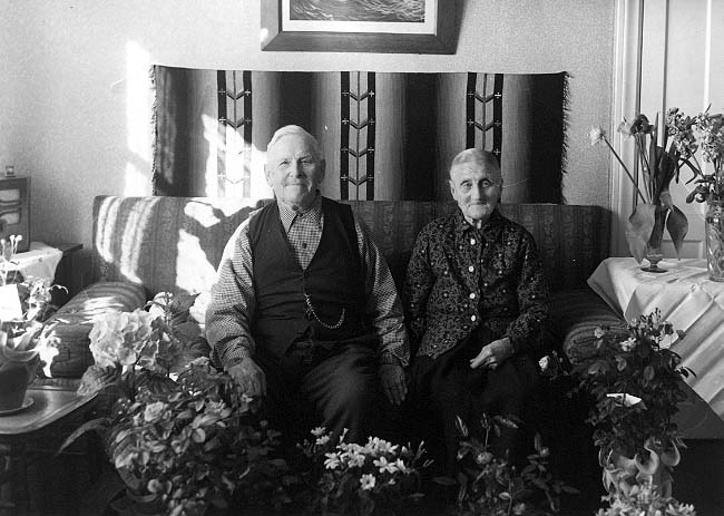 Ett äldre par sitter intill varandra i en soffa, de håller möjligen varandras hand. Solen strilar in från ett fönster till vänster. Det är paret Lätt som firar Järnbröllop.