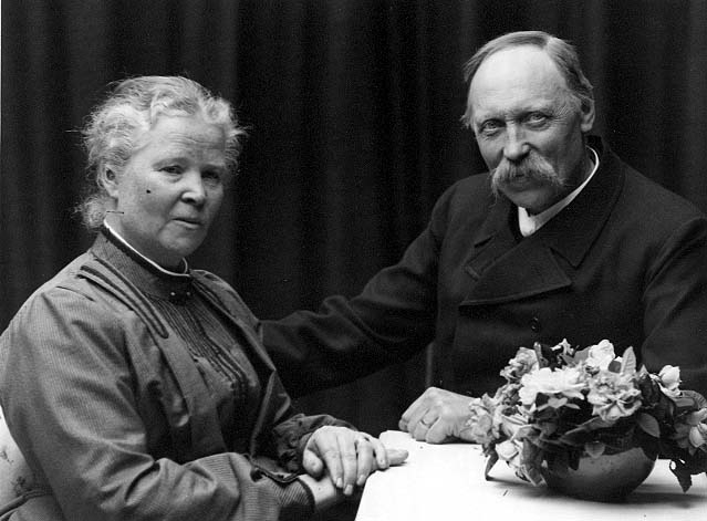 Gruppbild med det äldre paret Anna och C A Eklöf. Det sitter vid ett bord med en skål med blommor.